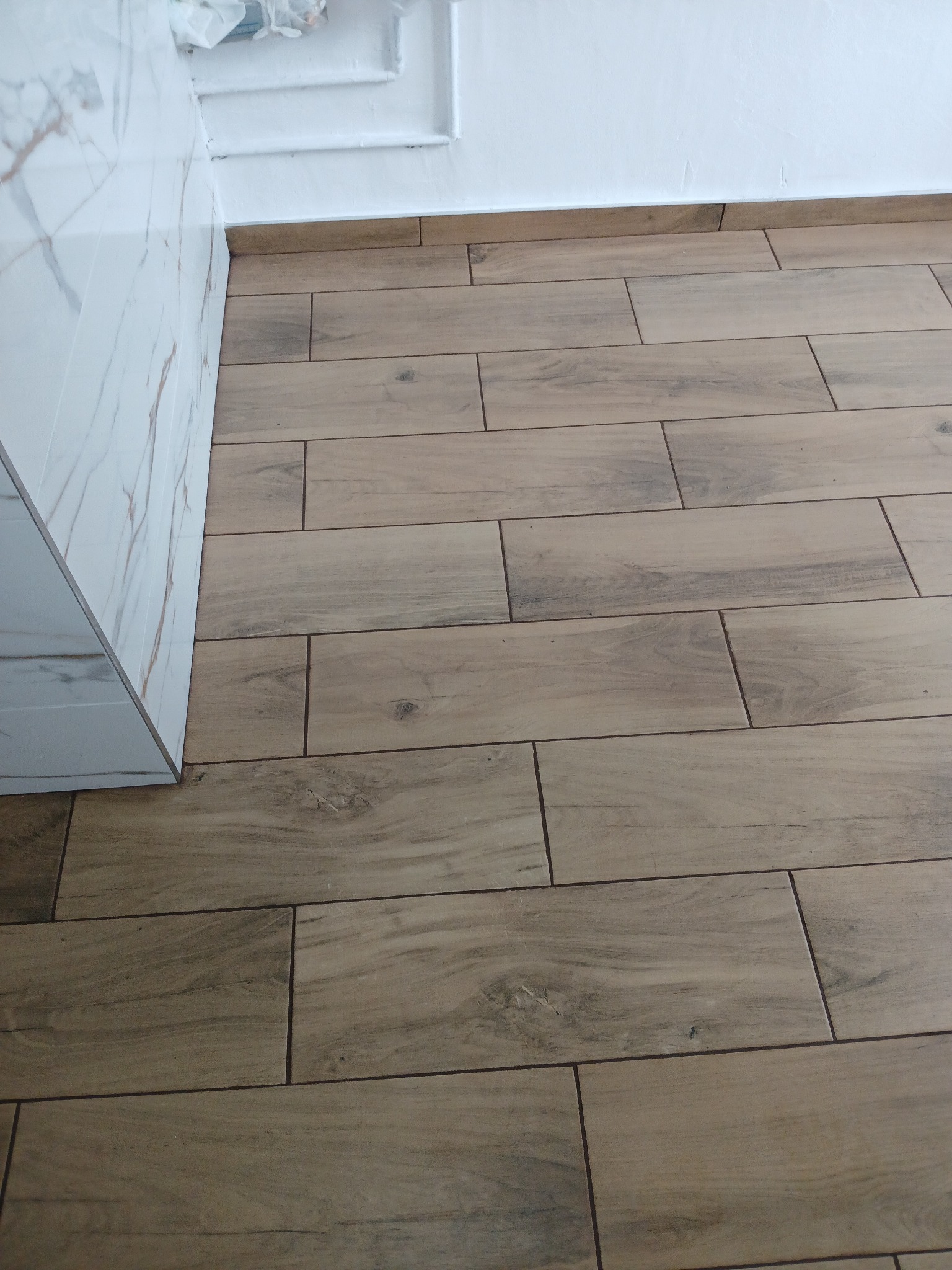 Ułożona podłoga z płytek imitujących drewno, widoczne fugi i fragment ściany wykończonej marmurowymi płytami oraz biały cokół.