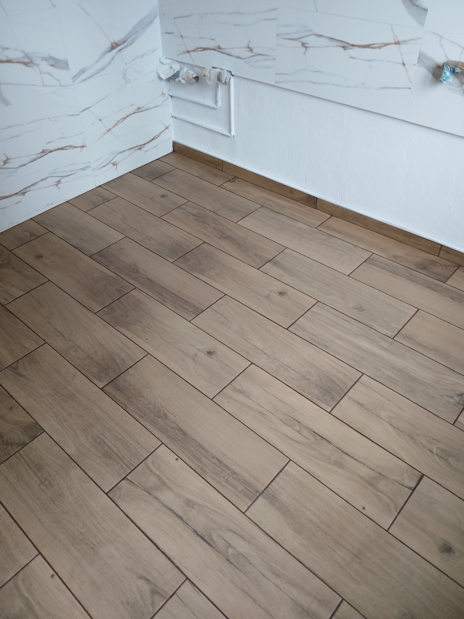 Ukończona podłoga z płytek imitujących drewno ułożonych w jodełkę w rogu pomieszczenia z wykończonymi ścianami z białych płytek imitujących marmur i gładką białą ścianą z wystającymi rurami.