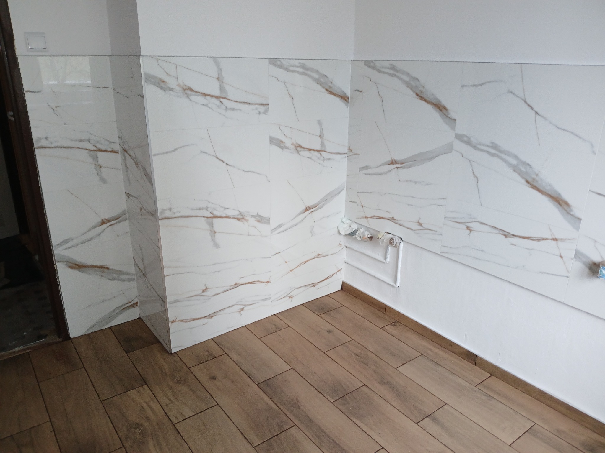 Wykończona łazienka z białymi płytkami imitującymi marmur z brązowymi żyłkami na ścianach i panelami podłogowymi w odcieniach brązu, widoczne rury hydrauliczne przy ścianie.