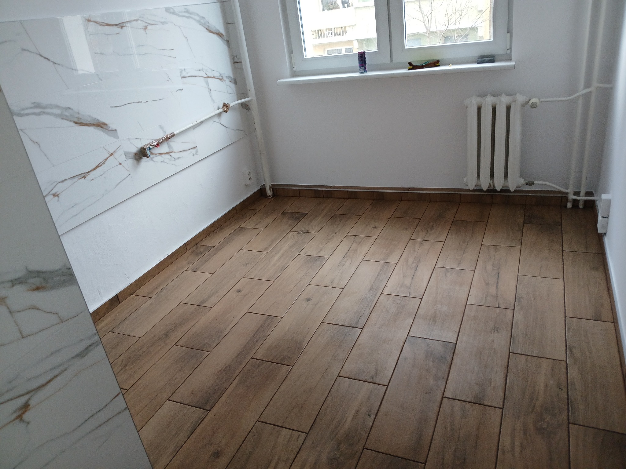 Wykończone wnętrze z drewnopodobną podłogą ułożoną w jodełkę, białą ścianą z płytami imitującymi marmur i grzejnikiem pod oknem.
