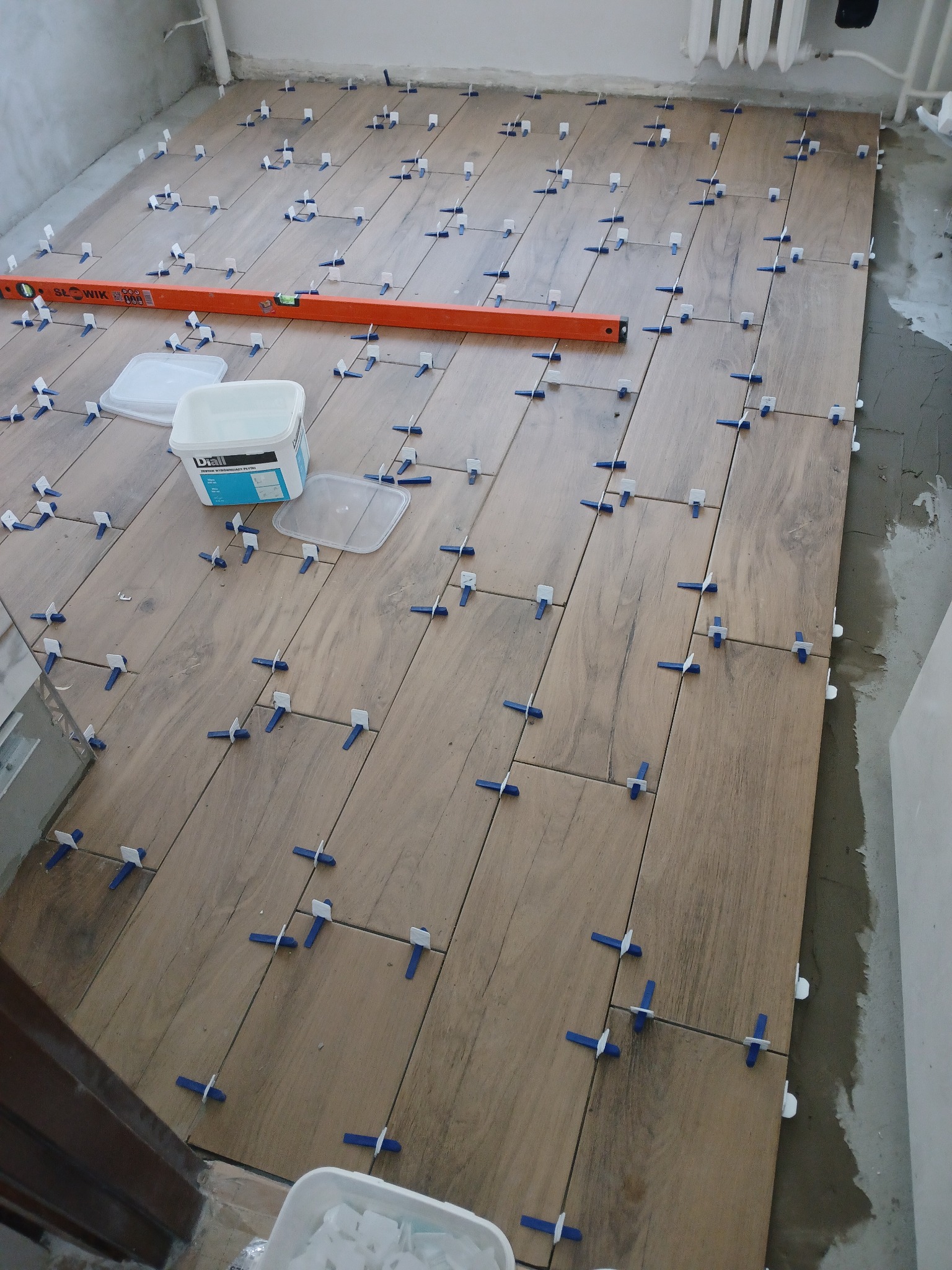 Układanie płytek podłogowych imitujących drewno, z użyciem klipsów poziomujących, widoczna poziomica i wiadro z klejem.