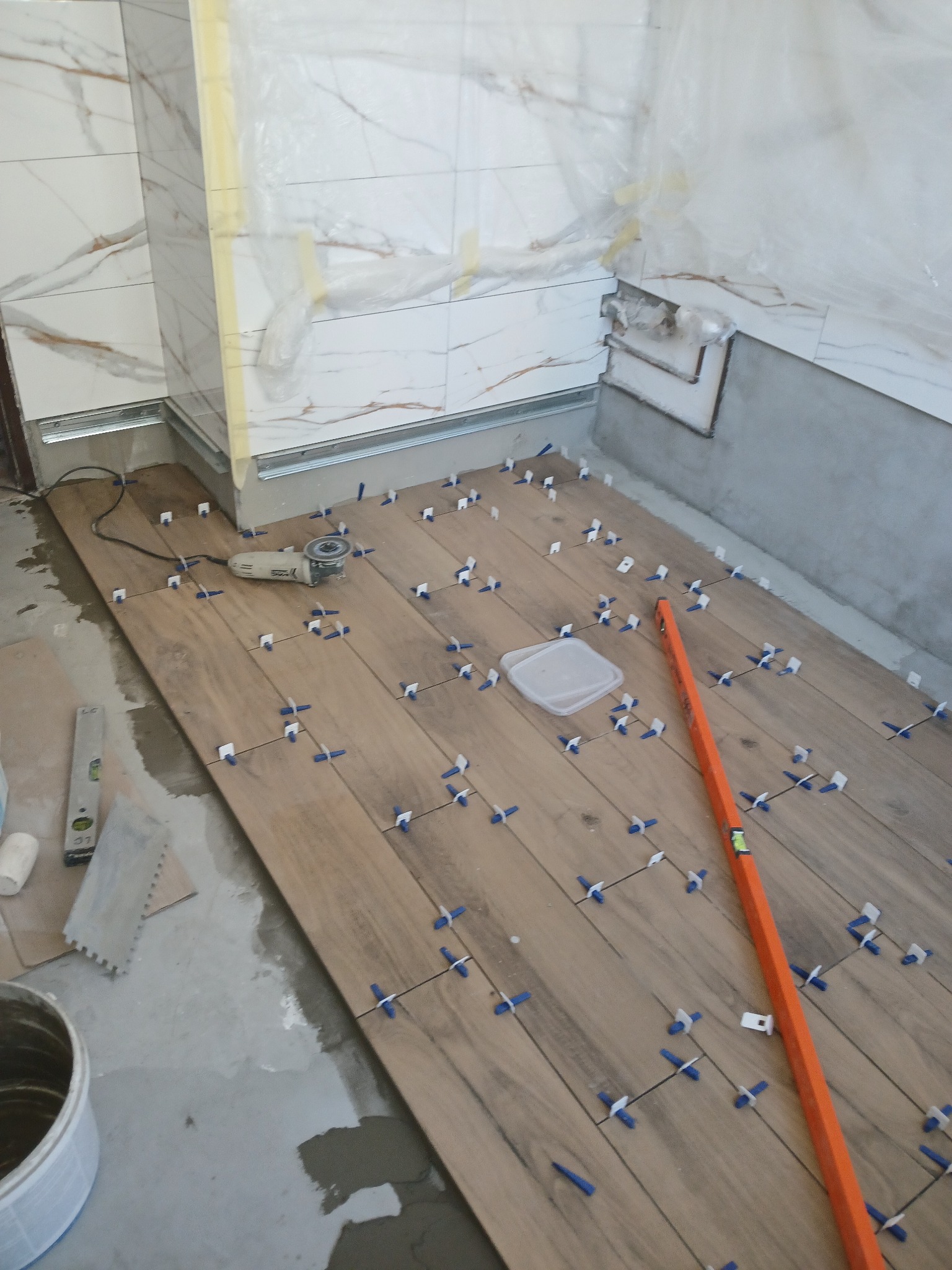 Układanie drewnopodobnych płytek podłogowych w łazience z białymi płytkami ściennymi, widoczne poziomice, szlifierka kątowa i krzyżyki dystansowe.