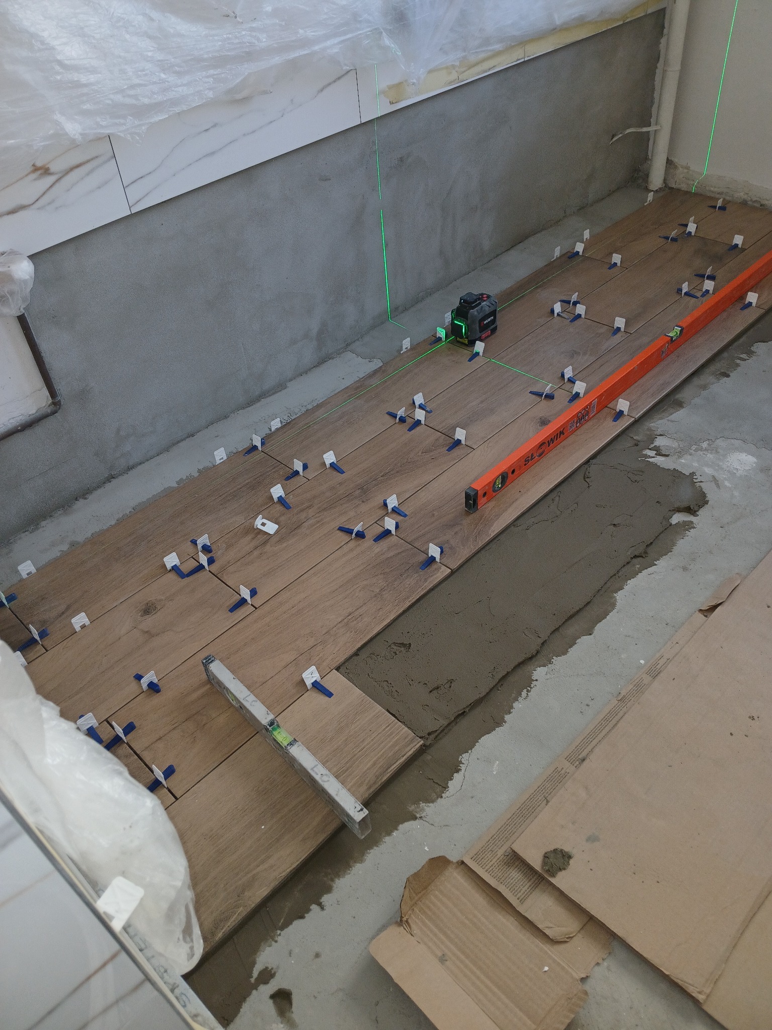 Układanie drewnopodobnych płytek podłogowych z użyciem poziomicy laserowej, poziomicy tradycyjnej i krzyżyków dystansowych; widoczny fragment ściany z ułożonymi białymi płytkami.