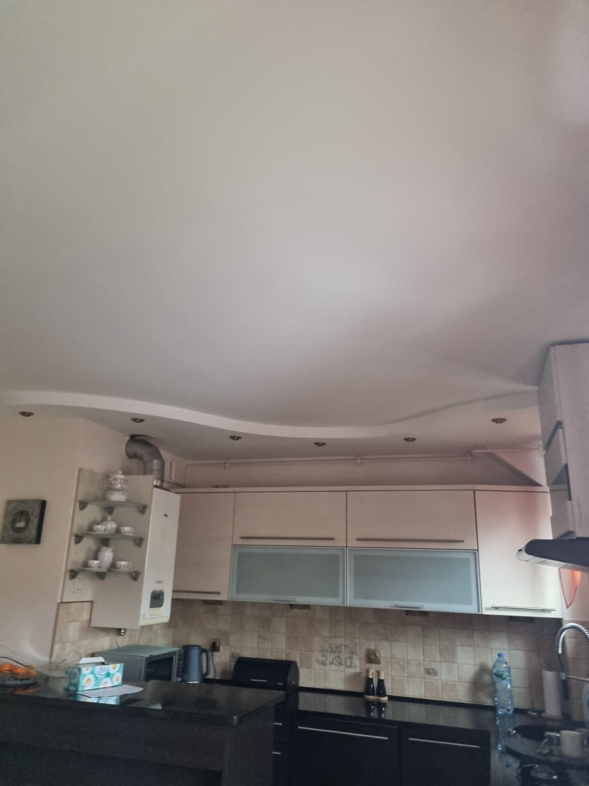 Kuchnia po remoncie: kremowe szafki wiszące z matowymi szybami, czarny blat, dekoracyjny sufit podwieszany z oświetleniem punktowym, oraz fragment ściany z półkami na porcelanę i piecem gazowym.