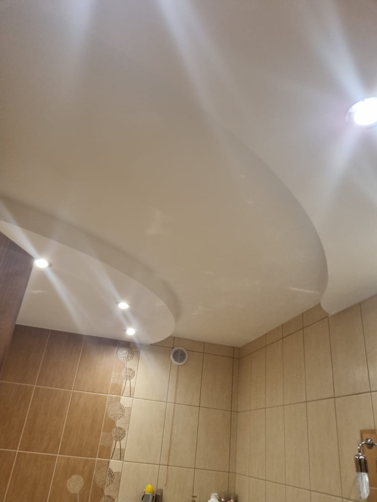 Zakrzywiony sufit podwieszany z wbudowanymi punktowymi lampami LED w łazience z brązowymi płytkami ceramicznymi z motywem dmuchawców.