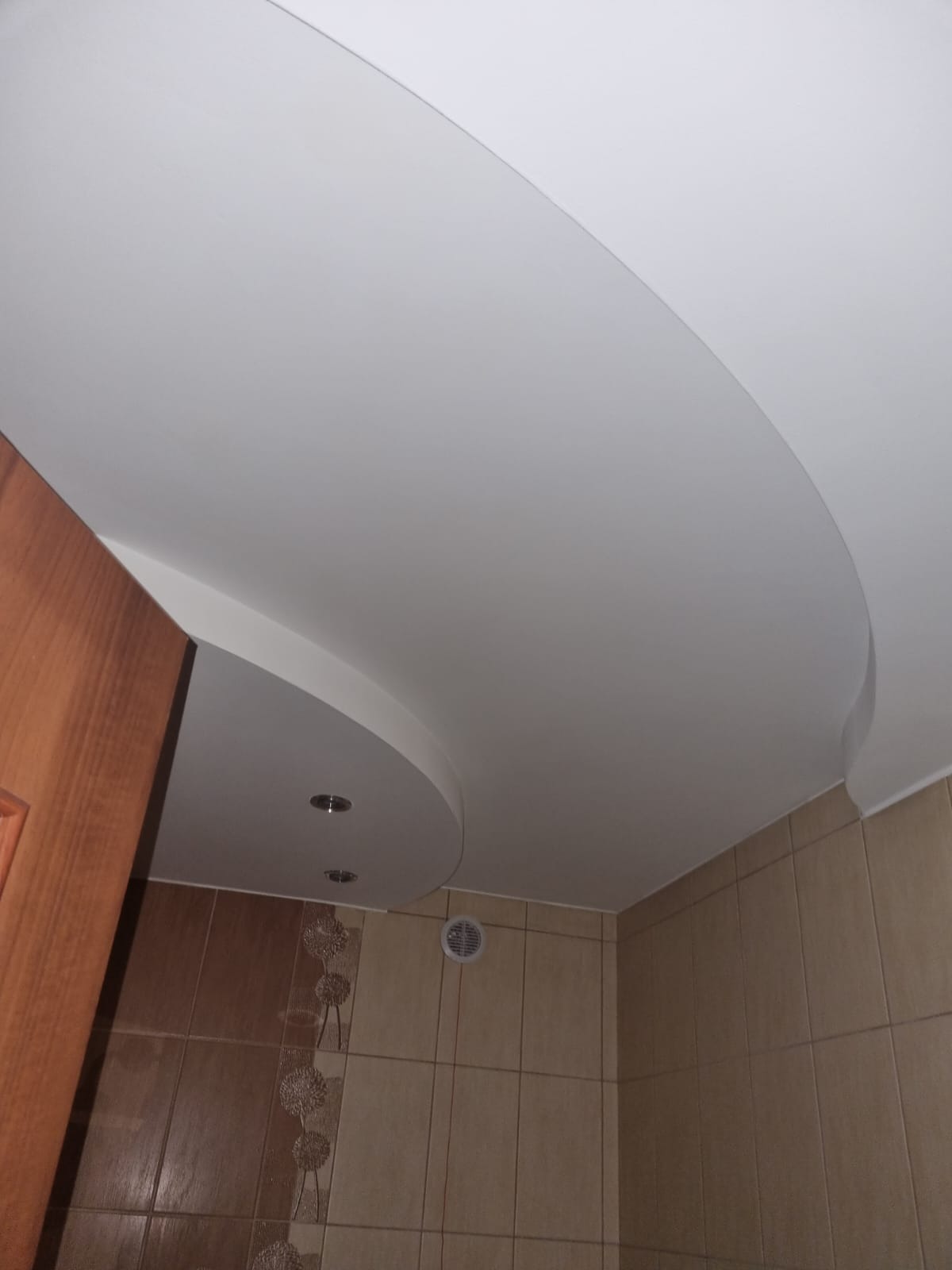 Podwieszany sufit z płyt kartonowo-gipsowych o nieregularnym kształcie z zamontowanymi punktowymi lampami halogenowymi w łazience z beżowymi płytkami.