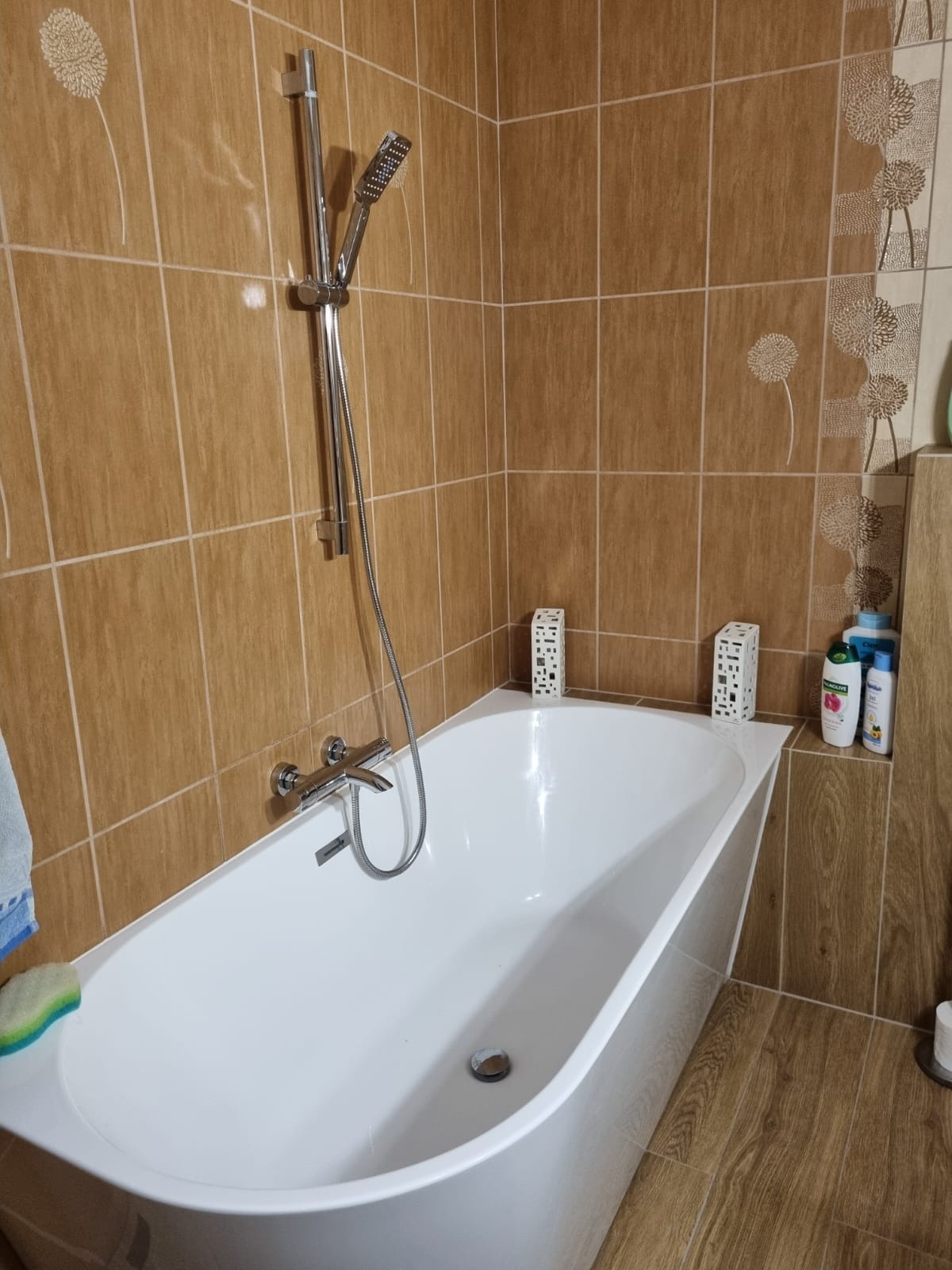 Biała wanna obudowana brązowymi płytkami z motywem dmuchawca, chromowana bateria i słuchawka prysznicowa, dekoracyjne ażurowe lampiony i kosmetyki na półce.