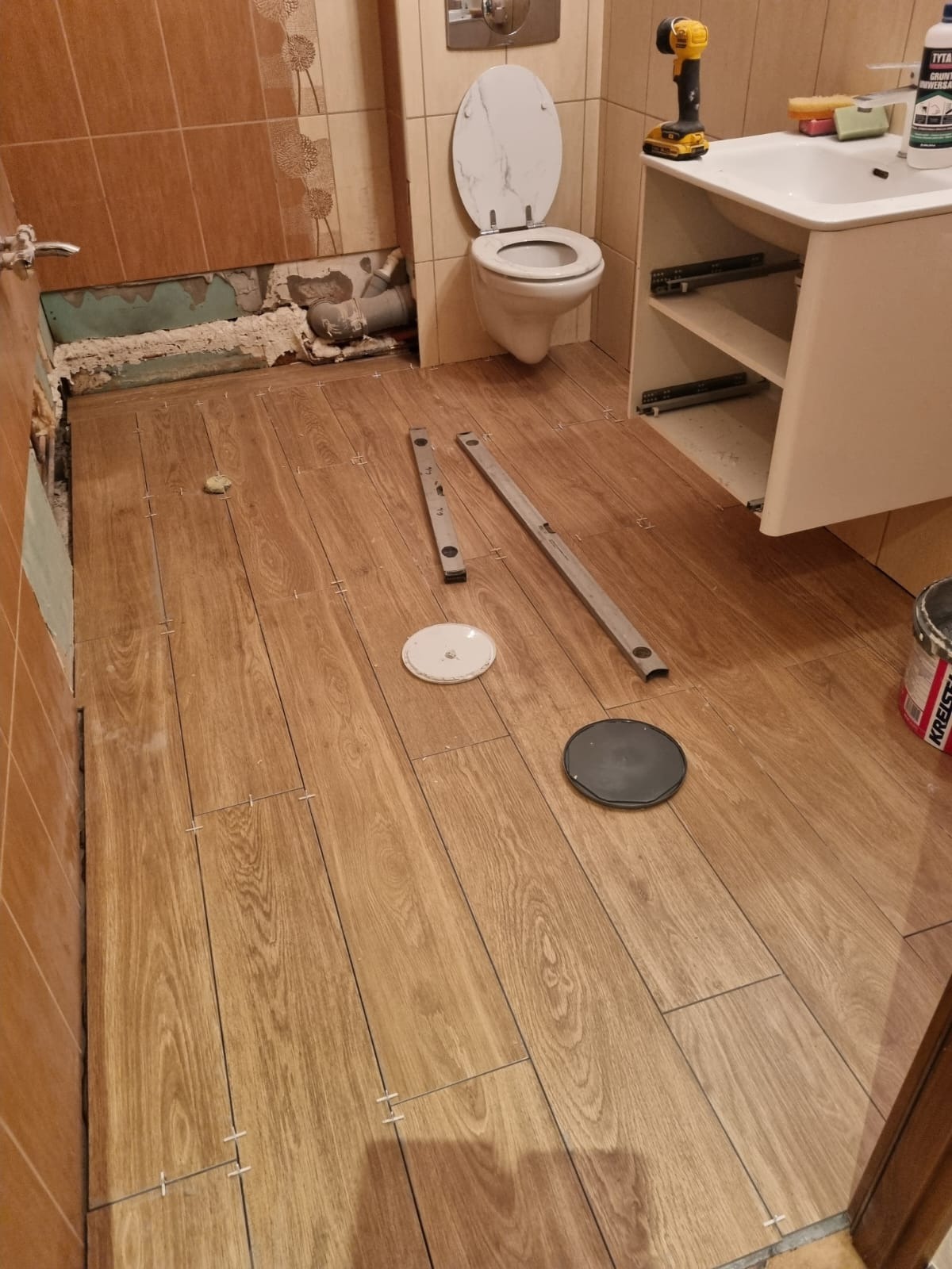 Podłoga w łazience w trakcie układania płytek imitujących drewno, widoczne poziomice, biała umywalka z szafką, toaleta i niedokończona ściana z instalacją.