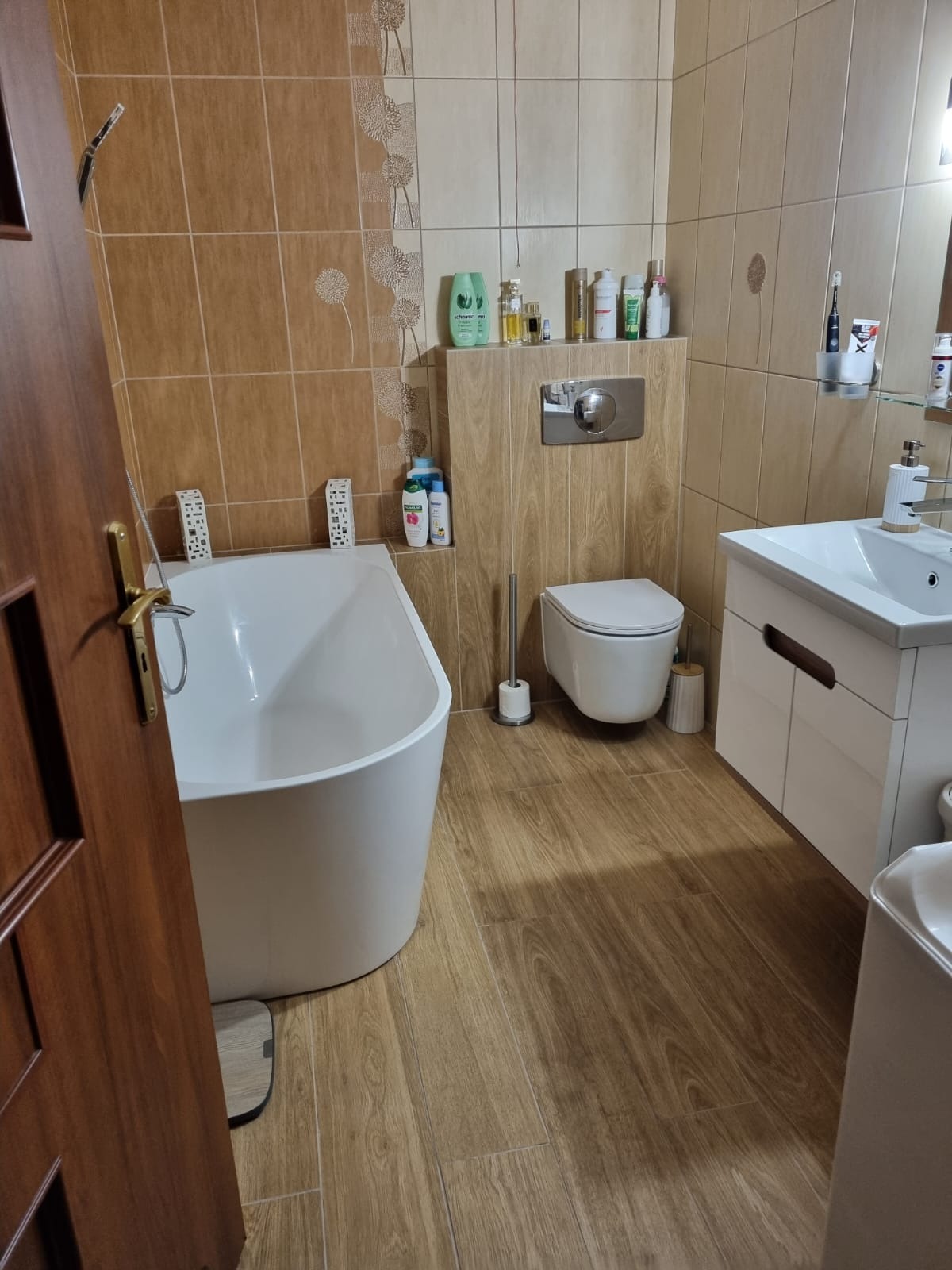 Wykończona łazienka w Wałbrzychu z brązowymi płytkami imitującymi drewno, białą wanną wolnostojącą, toaletą podwieszaną i umywalką z szafką, widok zza otwartych drzwi.