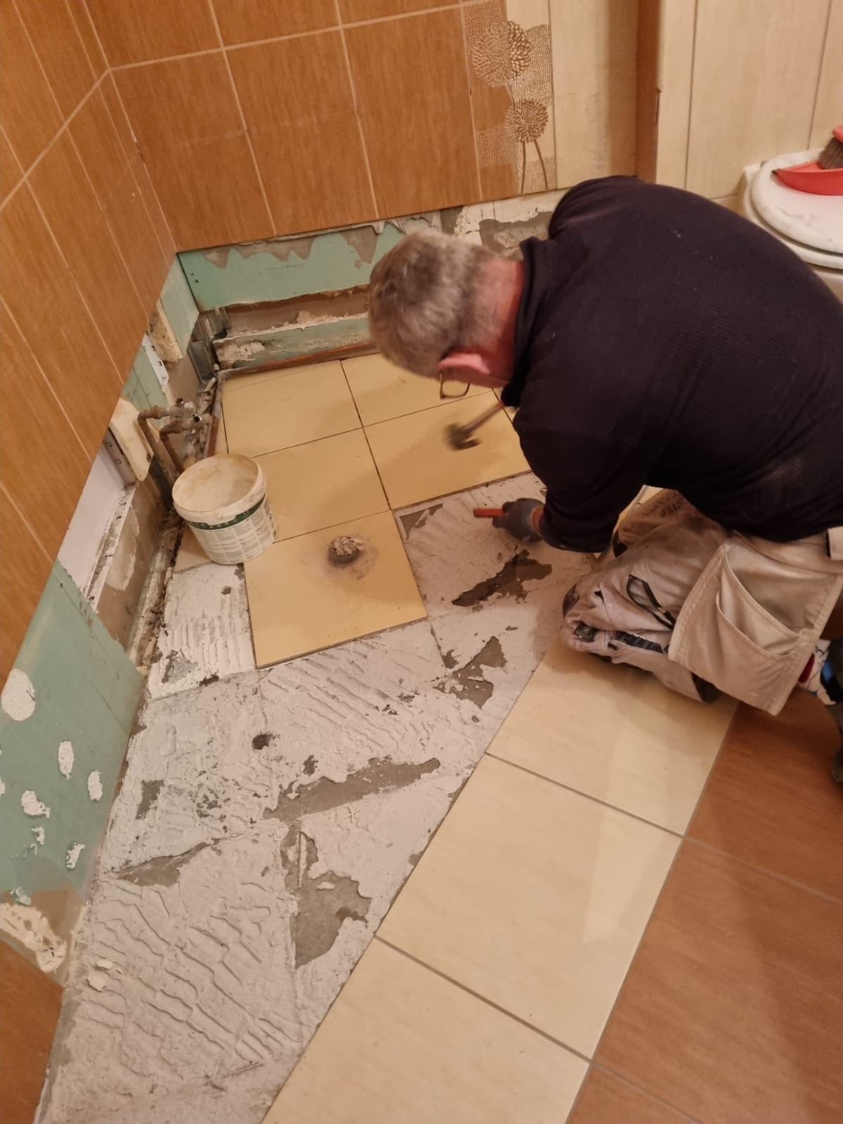 Mężczyzna w trakcie skuwania starych płytek podłogowych w łazience, widoczne fragmenty starej zaprawy i przygotowane nowe płytki.