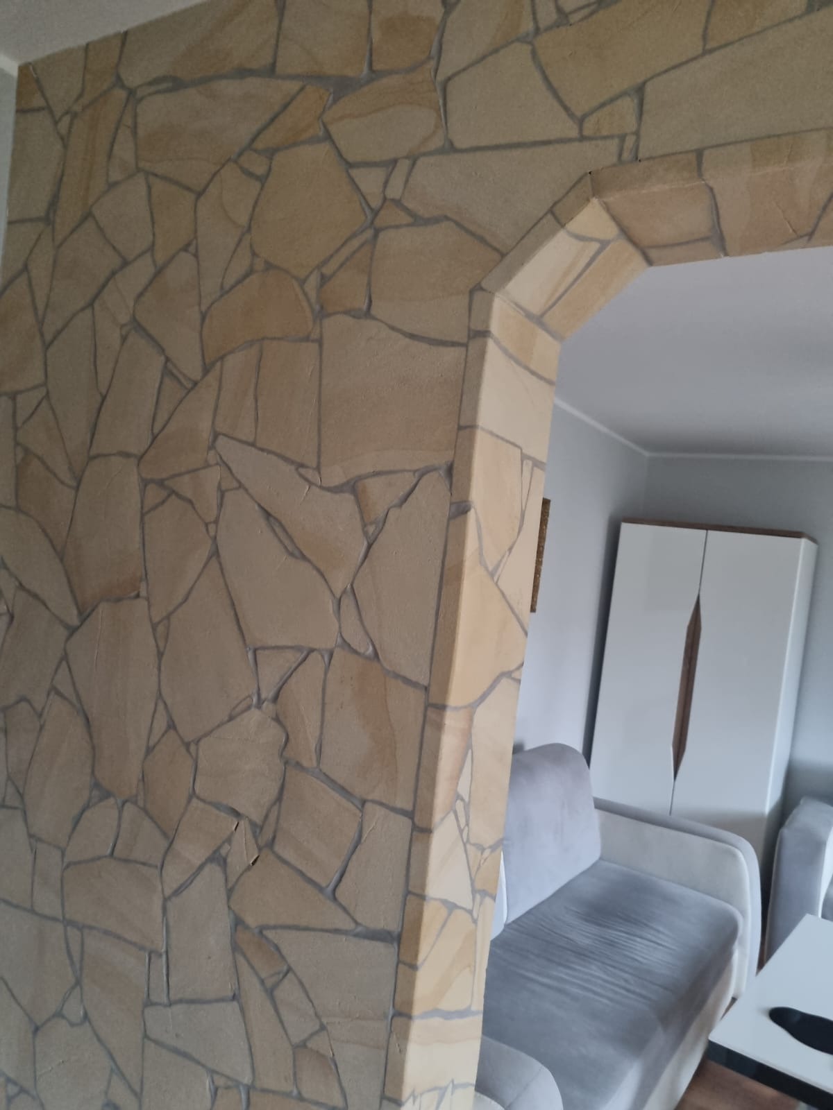 Ściana wykończona okładziną z piaskowca w kształcie nieregularnych kamieni, tworząca łuk wejściowy do pokoju z szarą sofą i białą szafą.