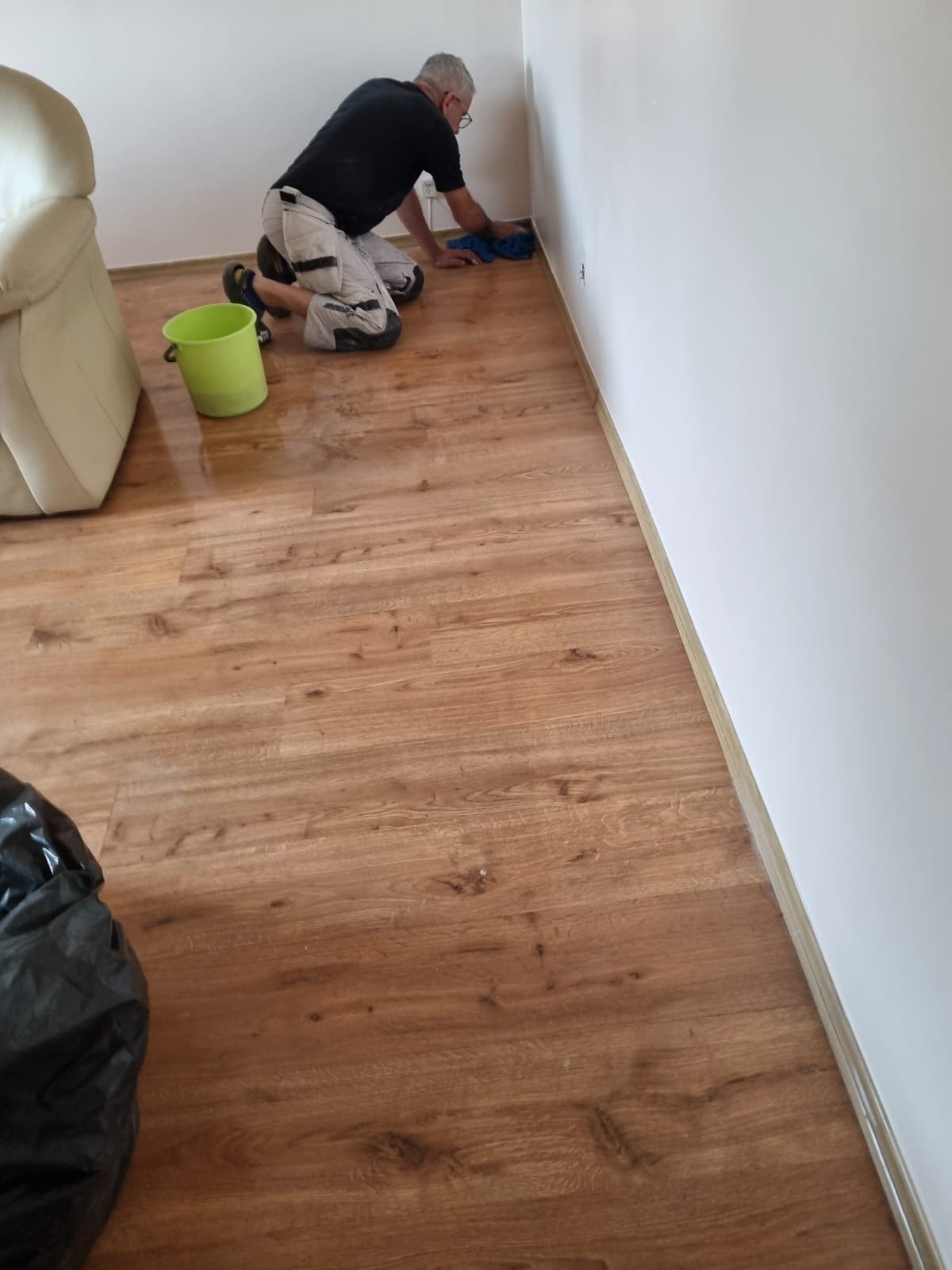 Mężczyzna na kolanach czyści ścianę w rogu pomieszczenia, widoczna podłoga z paneli, wiadro i fragment sofy.