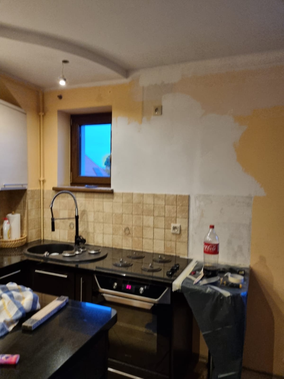 Kuchnia w trakcie odświeżania: ściana częściowo pokryta gładzią, widoczny fragment kuchenki, okno z widokiem na zewnątrz, butelka Coca-Coli na blacie.