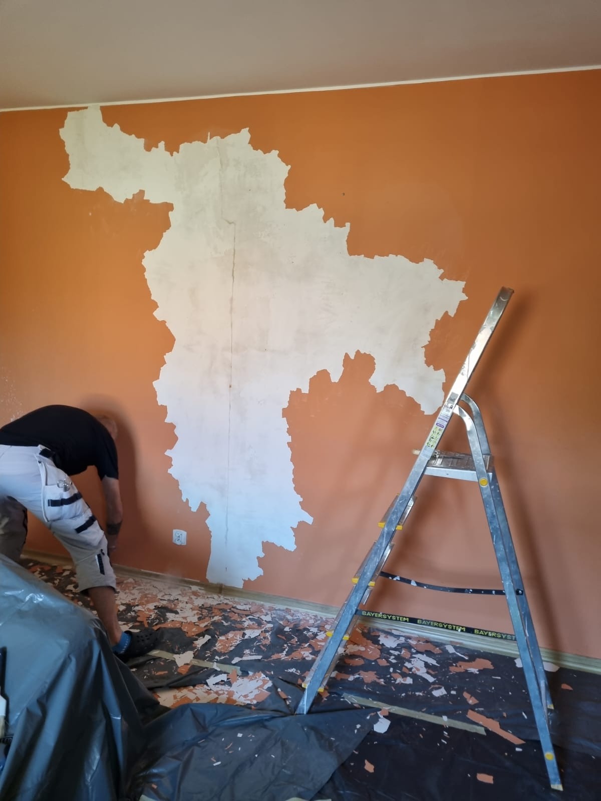Pracownik w trakcie usuwania starej farby ze ściany, widoczne fragmenty odspojonej farby na podłodze zabezpieczonej folią, drabina oparta o ścianę, ściana częściowo pokryta nową warstwą.