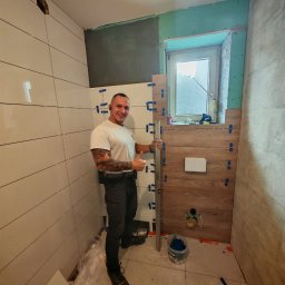 Jarosław Grądecki - Uśmiechnięty mężczyzna z tatuażami na ramionach, ubrany w białą koszulkę i ciemne spodnie, trzyma poziomicę i kciukiem w górę na tle częściowo wyłożonej kafelkami łazienki z elementami imitującymi...