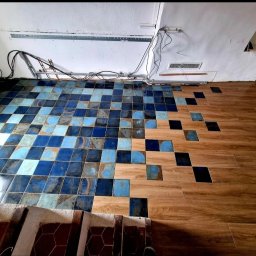 Jarosław Grądecki - Nietypowe połączenie płytek ceramicznych w odcieniach niebieskiego z drewnopodobnymi panelami, tworzące efekt pikselizacji na podłodze w trakcie remontu.