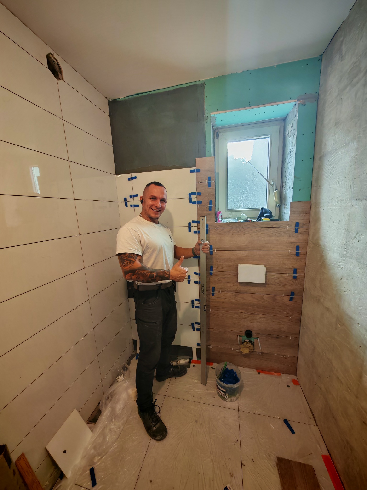 Uśmiechnięty mężczyzna z tatuażami na ramionach, ubrany w białą koszulkę i ciemne spodnie, trzyma poziomicę i kciukiem w górę na tle częściowo wyłożonej kafelkami łazienki z elementami imitującymi...