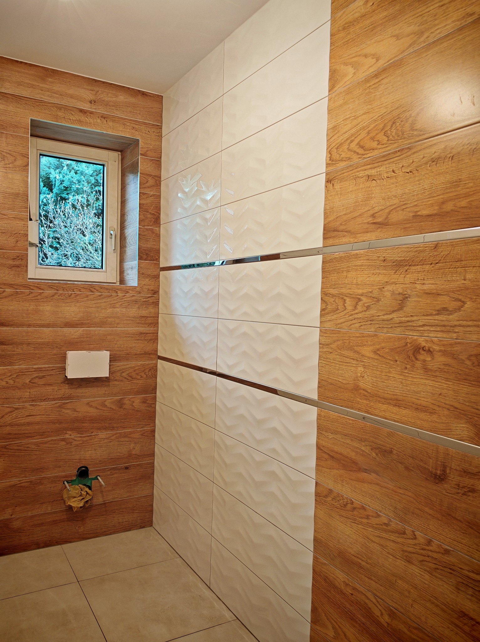 Nowoczesna łazienka z drewnopodobnymi panelami na ścianie, przechodzącymi w białe, teksturowane płytki oddzielone metalowymi listwami; widoczne okno z widokiem na drzewa.