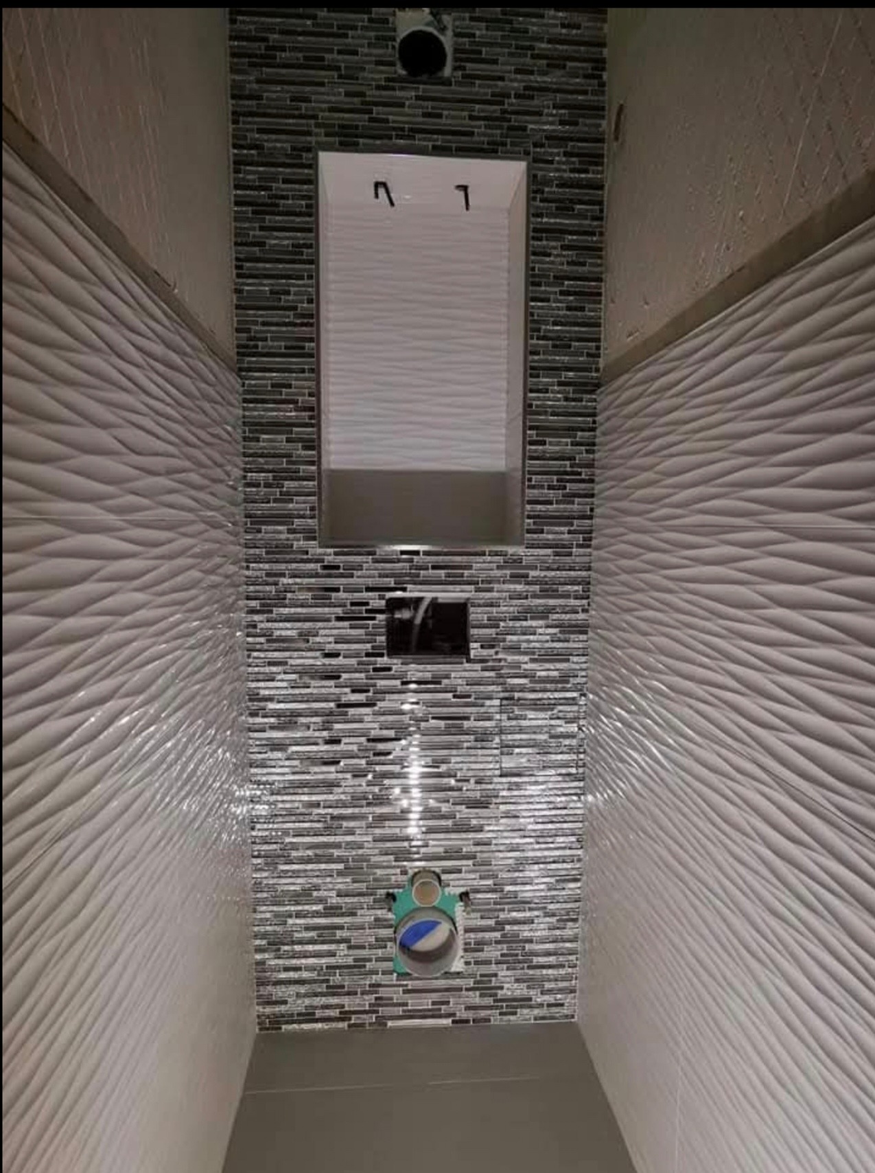 Wąska toaleta w trakcie wykańczania, ściany wyłożone białą, falistą glazurą oraz mozaiką w odcieniach szarości, widoczne przyłącze do toalety podtynkowej.