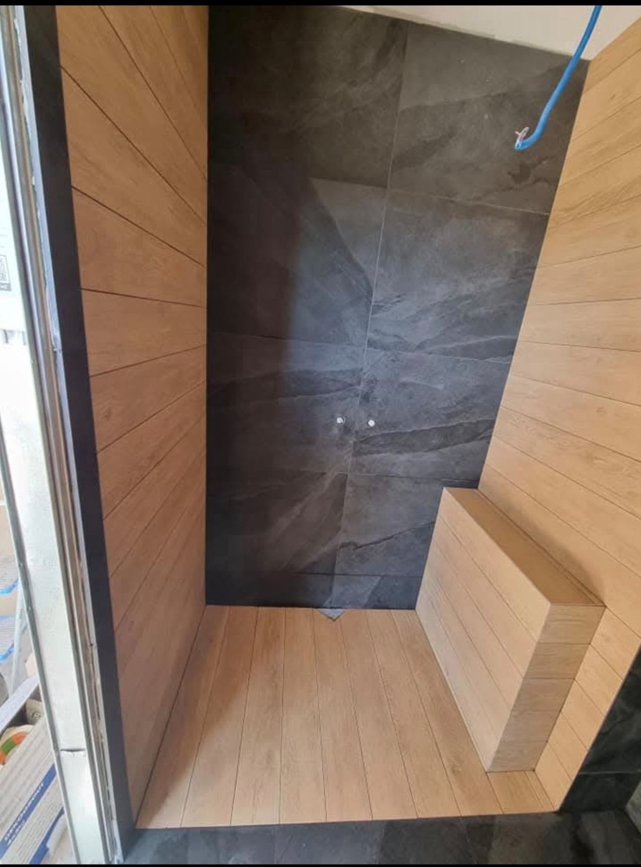 Nowoczesna łazienka w trakcie wykańczania z drewnopodobnymi płytkami na ścianach i podłodze, kontrastującymi z szarą, kamienną fakturą płytek pod prysznicem; widoczny wystający kabel elektryczny.