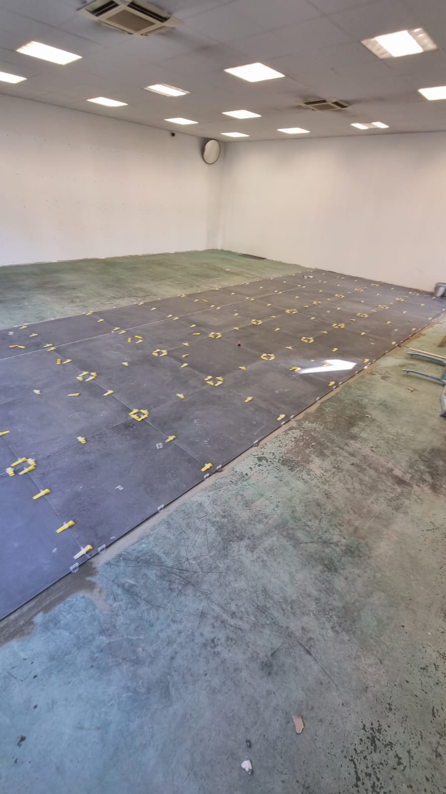 Przygotowanie do fugowania: Ułożone ciemnoszare płytki podłogowe z żółtymi krzyżykami dystansowymi na podłożu wymagającym wyrównania, widoczne jasne ściany i sufit z oświetleniem rastrowym.