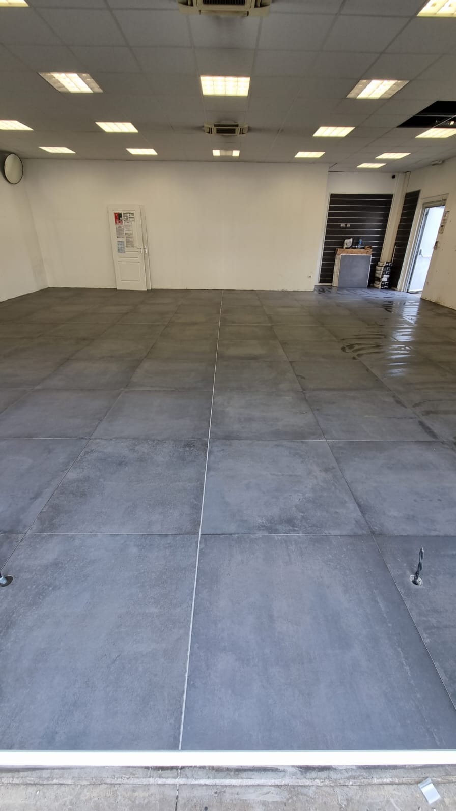 Nowo ułożona, duża, szara płytka podłogowa w przestronnym, jasnym pomieszczeniu z białymi ścianami i sufitem kasetonowym. Widoczne fugi i dwa otwory z wystającymi kablami.