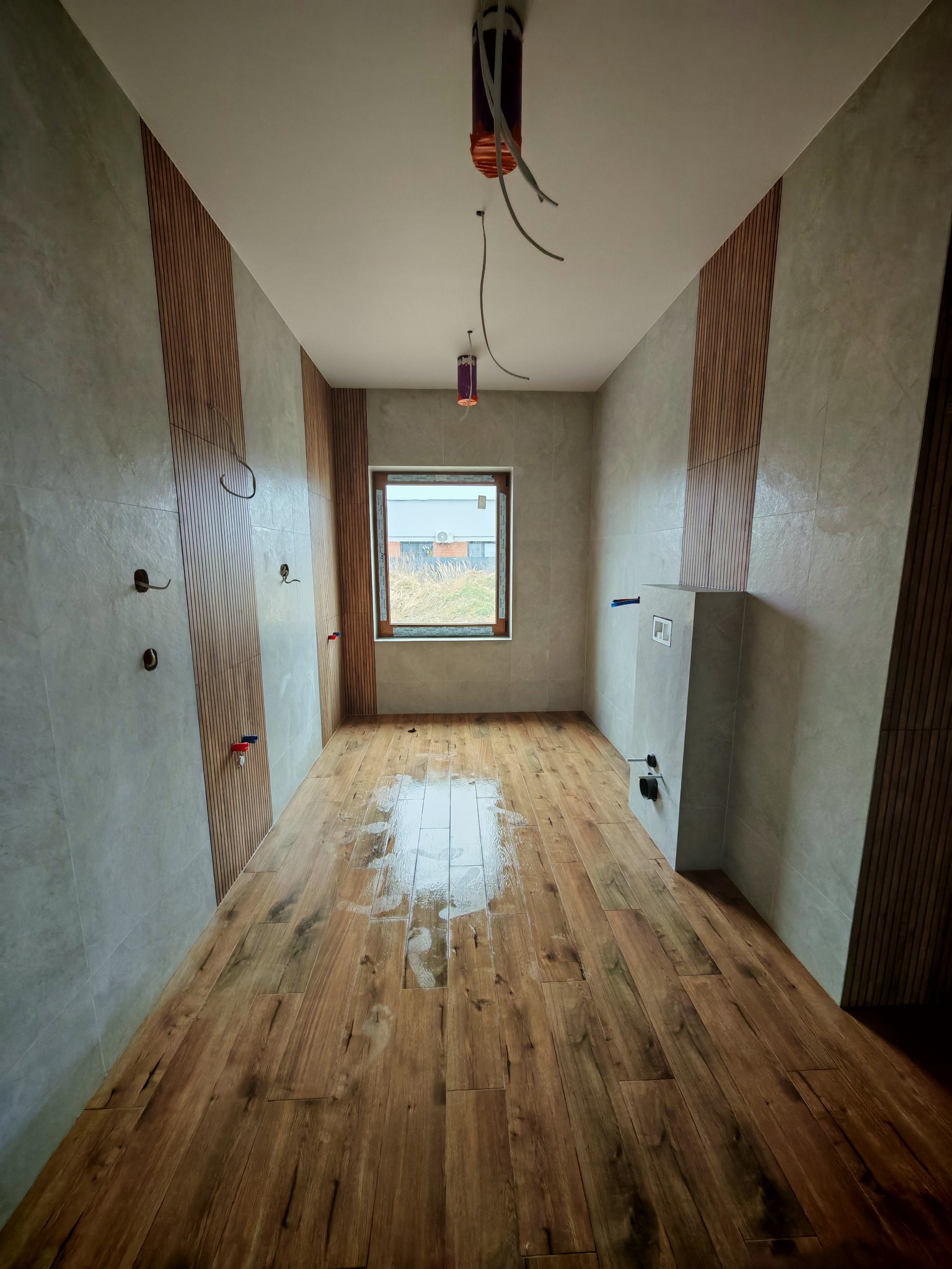 Wnętrze łazienki w trakcie prac wykończeniowych; widoczne drewnopodobne panele na podłodze, fragment ściany z szarymi płytkami i drewnianymi panelami dekoracyjnymi, instalacja podtynkowa WC...