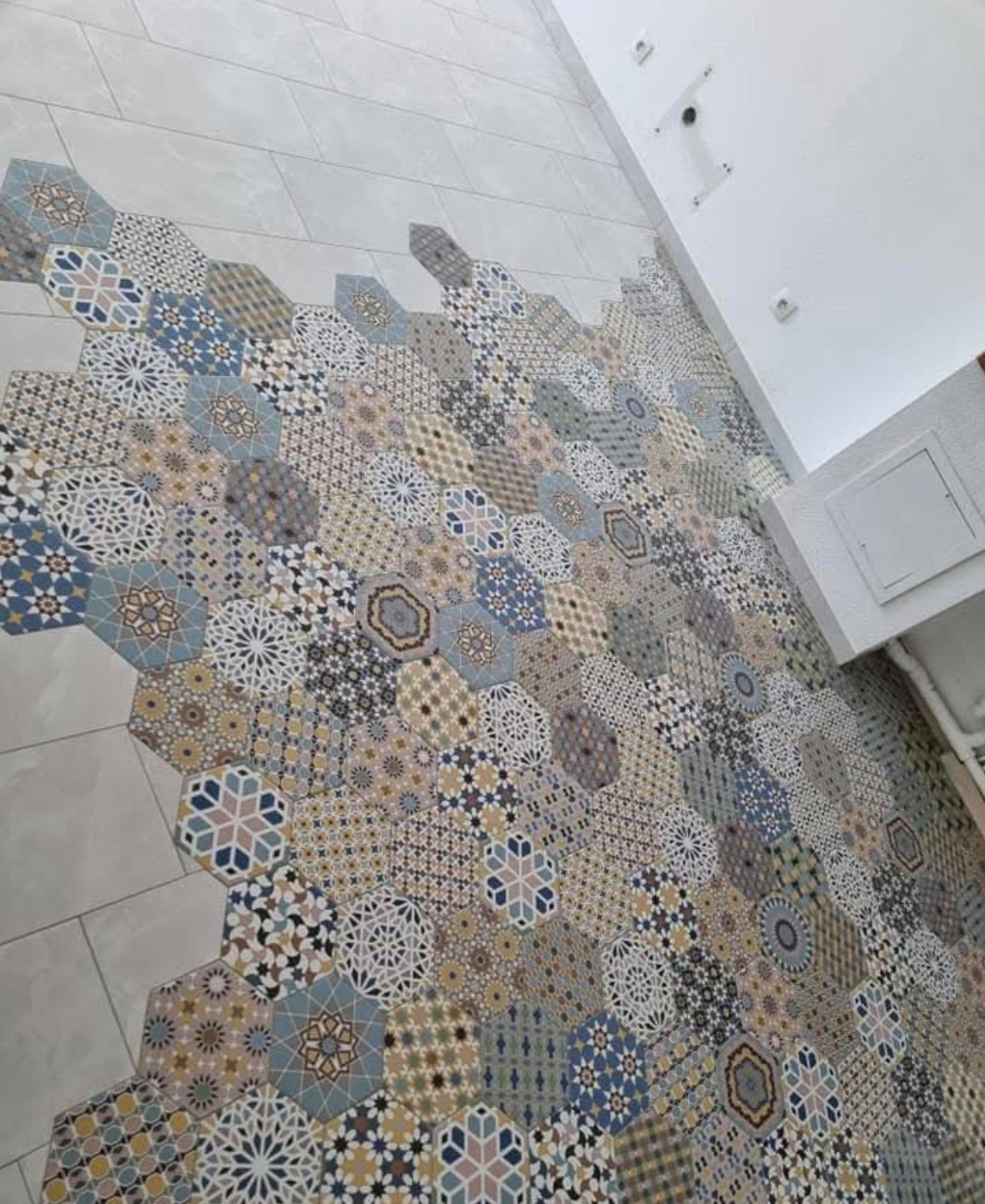 Ściana wykończona dekoracyjnymi, heksagonalnymi płytkami w różnych wzorach i kolorach, przechodząca w gładką, jasną ścianę. Widoczne elementy instalacji elektrycznej i fragment białej szafki.