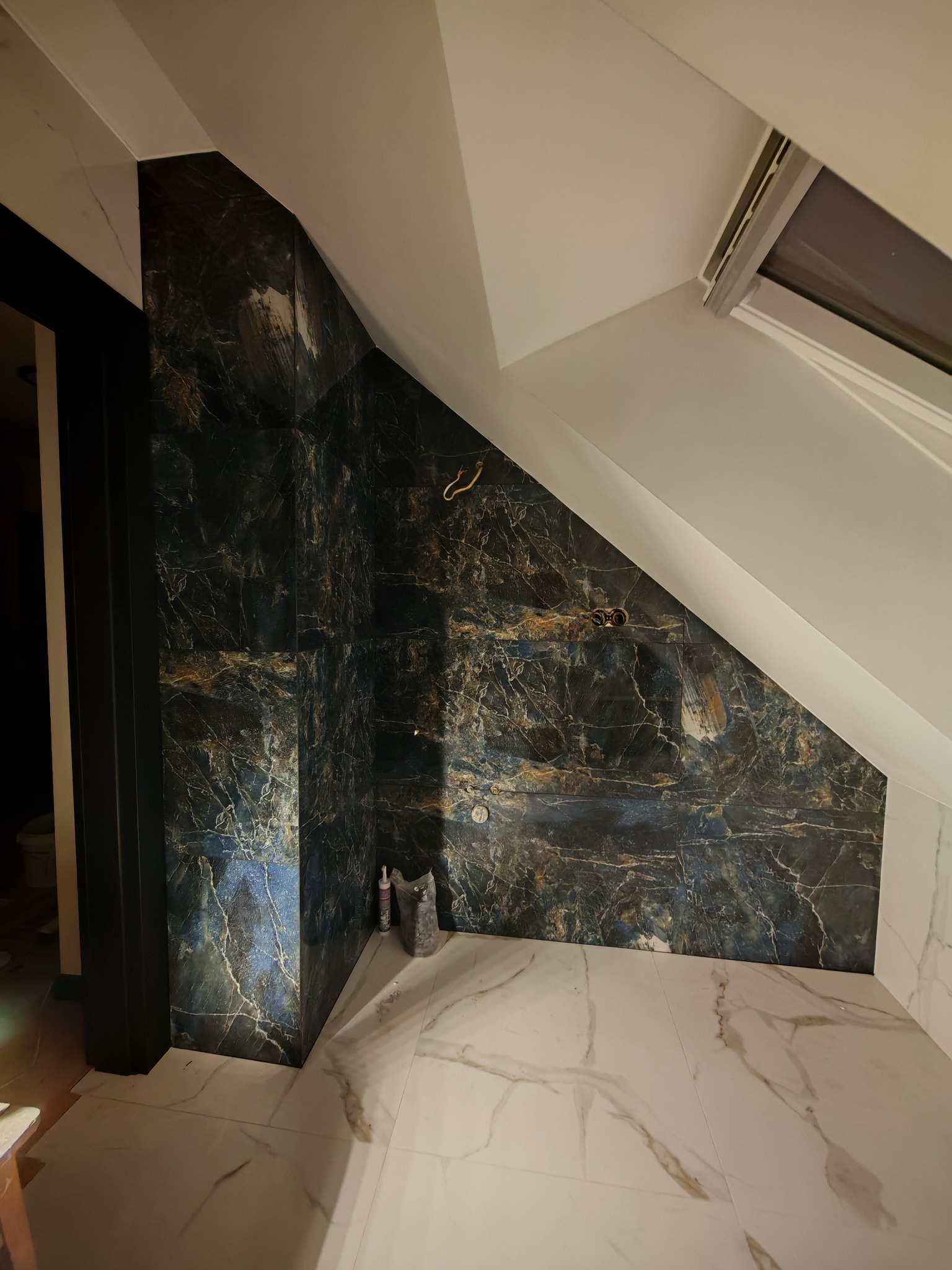 Wykończona łazienka na poddaszu z marmurowymi płytkami w odcieniach bieli i granatu. Widoczne gniazdka elektryczne i okno dachowe.