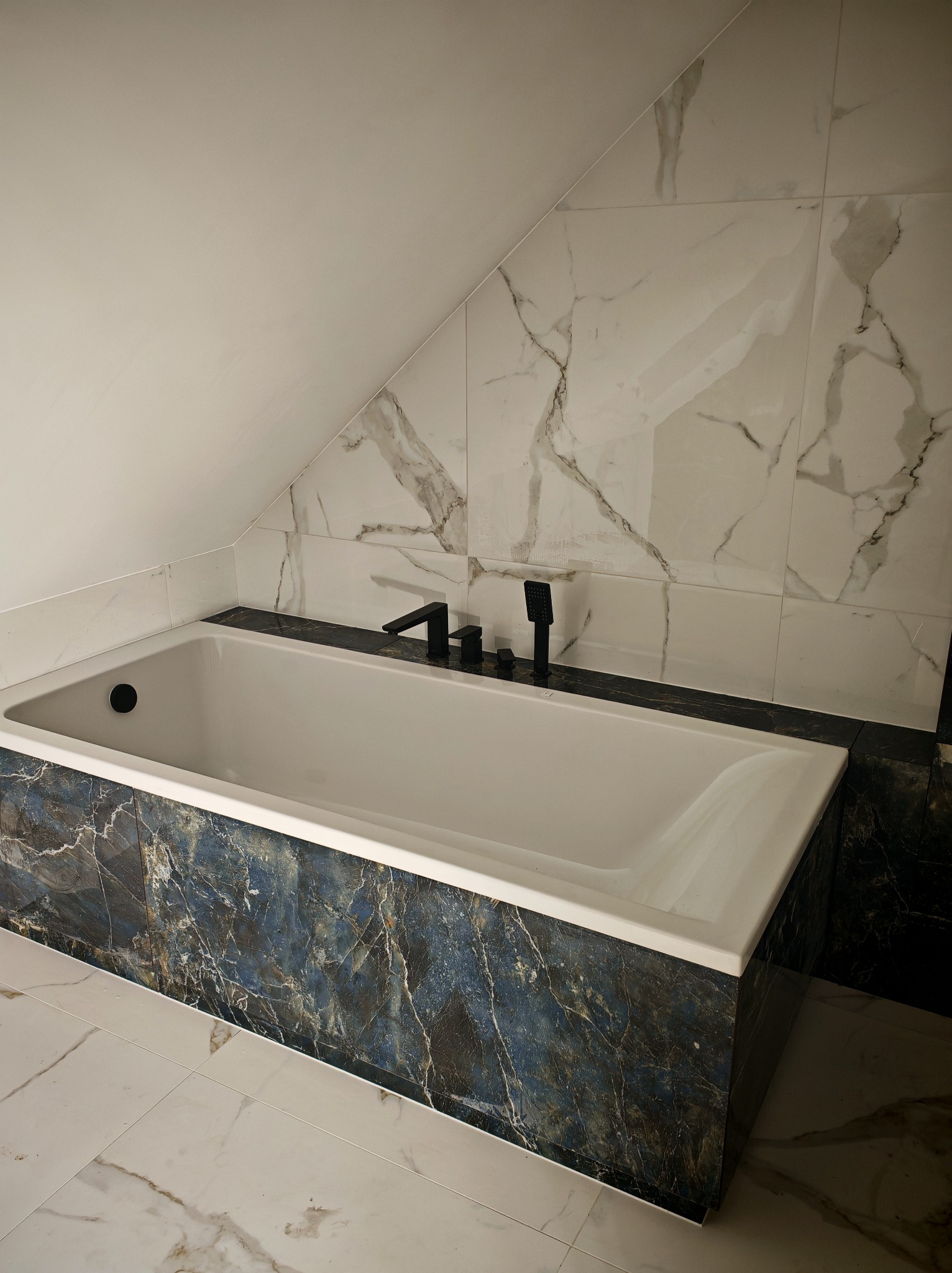 Biała wanna w obudowie z ciemnego marmuru, czarna armatura, ściany i podłoga wyłożone płytkami imitującymi biały marmur z szarymi żyłami, skośny sufit.