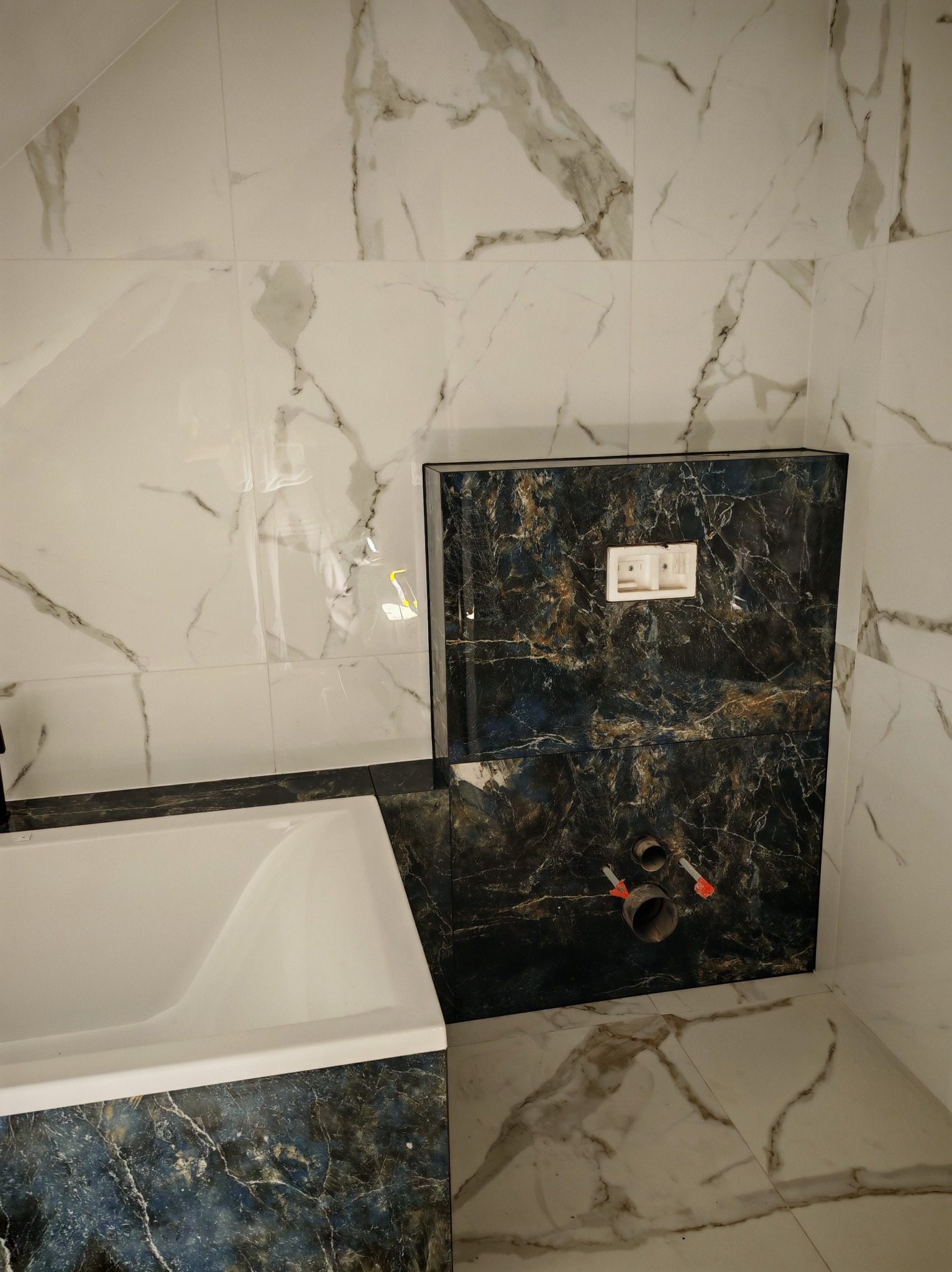 Wykończona łazienka z wanną i zabudową WC w marmurze. Białe płytki z szarymi żyłami na ścianach i podłodze, ciemny marmur na obudowie wanny i stelażu WC.