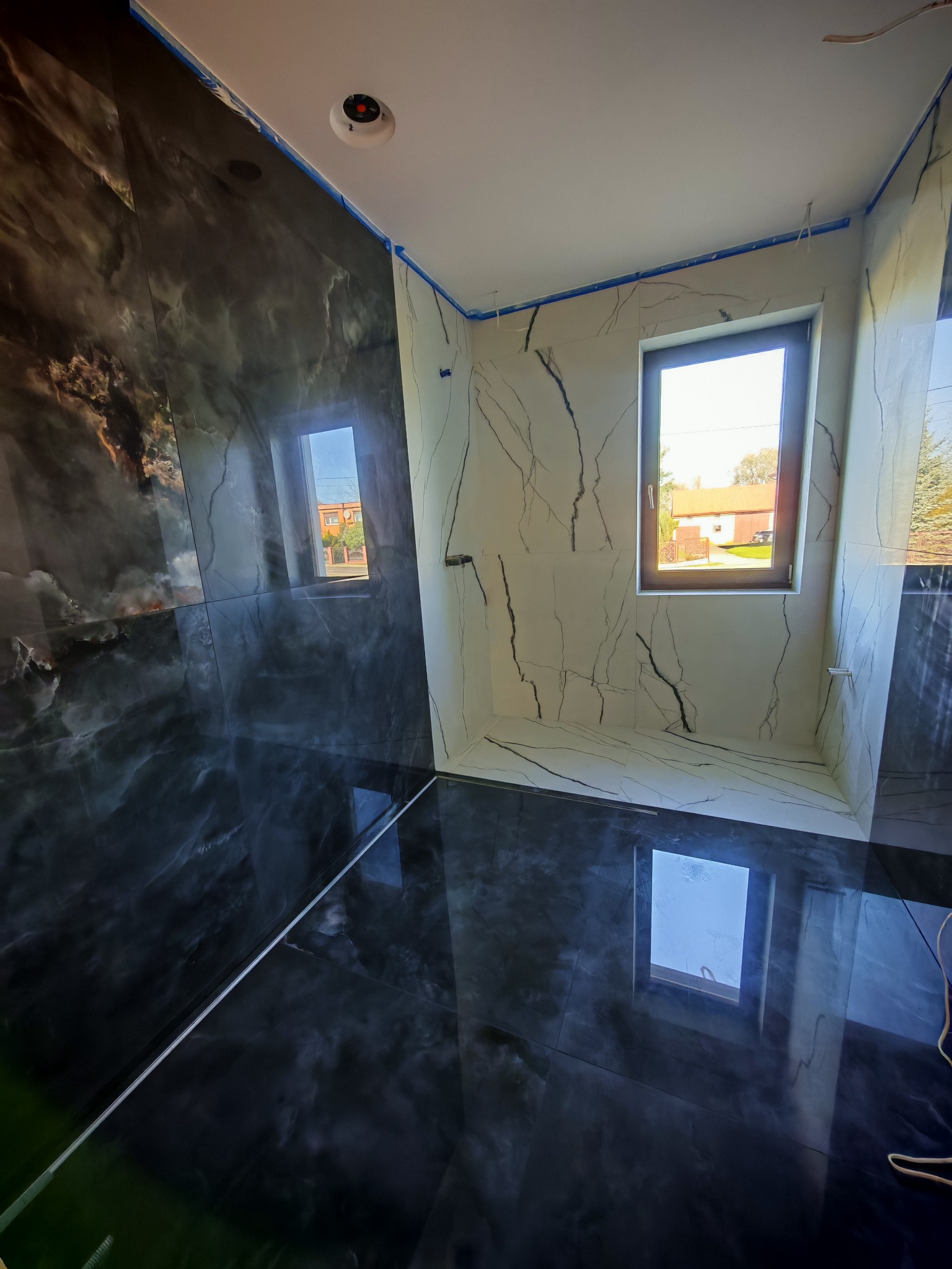 Nowoczesna łazienka z czarno-białymi płytkami imitującymi marmur, okno z widokiem na zewnątrz. Widoczne taśmy malarskie zabezpieczające krawędzie.