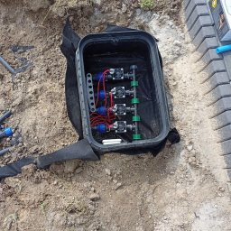 JAROSŁAW KOŚMICKI - Skrzynka zaworowa z rozdzielaczem i elektrozaworami w trakcie montażu w ziemi, widoczne przewody i elementy instalacji nawadniającej oraz fragment ułożonej kostki brukowej.
