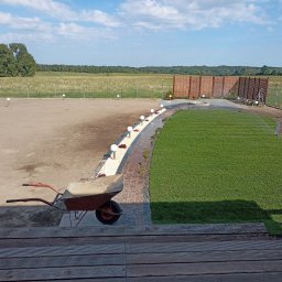 JAROSŁAW KOŚMICKI - Widok z góry na teren po częściowym ułożeniu trawy z rolki, z widoczną taczką na pierwszym planie, ścieżką z białego żwiru i kulistymi lampami, oraz drewnianym ogrodzeniem panelowym w tle.