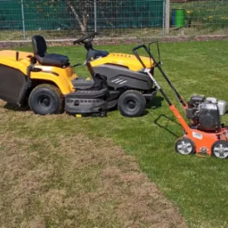 Żółty traktorek do koszenia trawy i wertykulator na trawniku, z widoczną różnicą w wyglądzie trawy po zabiegu wertykulacji.