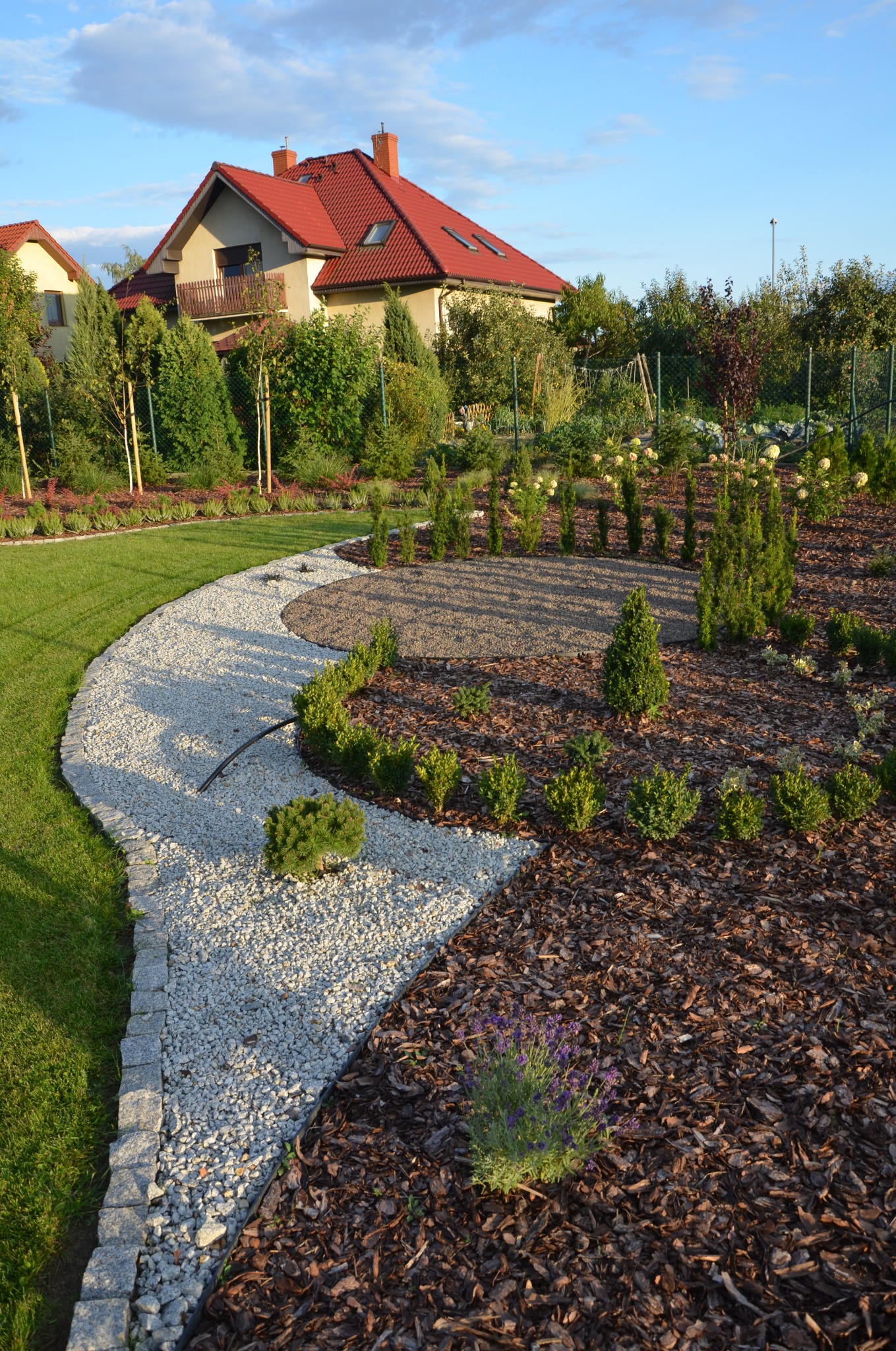 Widok na nowo założony ogród z elementami dekoracyjnymi: ścieżka z białego żwiru obramowana kostką, rabaty wysypane korą, świeżo posadzone krzewy i drzewka iglaste, trawnik oraz dom z czerwonym...