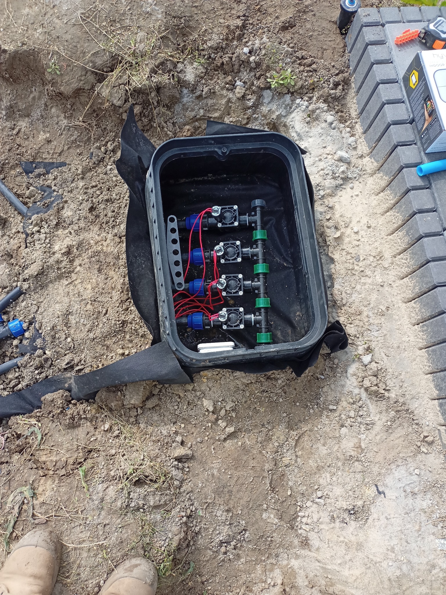 Skrzynka zaworowa z rozdzielaczem i elektrozaworami w trakcie montażu w ziemi, widoczne przewody i elementy instalacji nawadniającej oraz fragment ułożonej kostki brukowej.