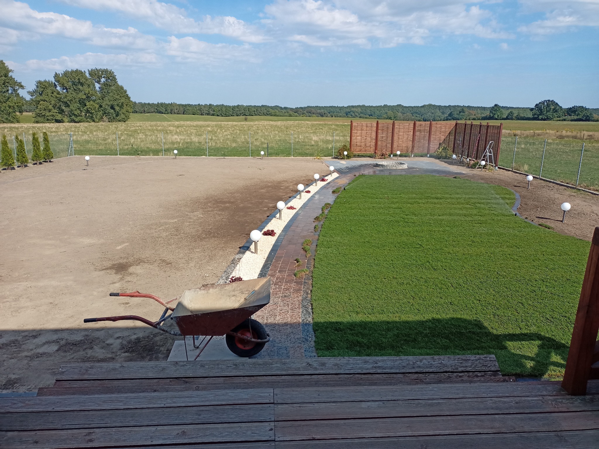 Widok z góry na teren po częściowym ułożeniu trawy z rolki, z widoczną taczką na pierwszym planie, ścieżką z białego żwiru i kulistymi lampami, oraz drewnianym ogrodzeniem panelowym w tle.
