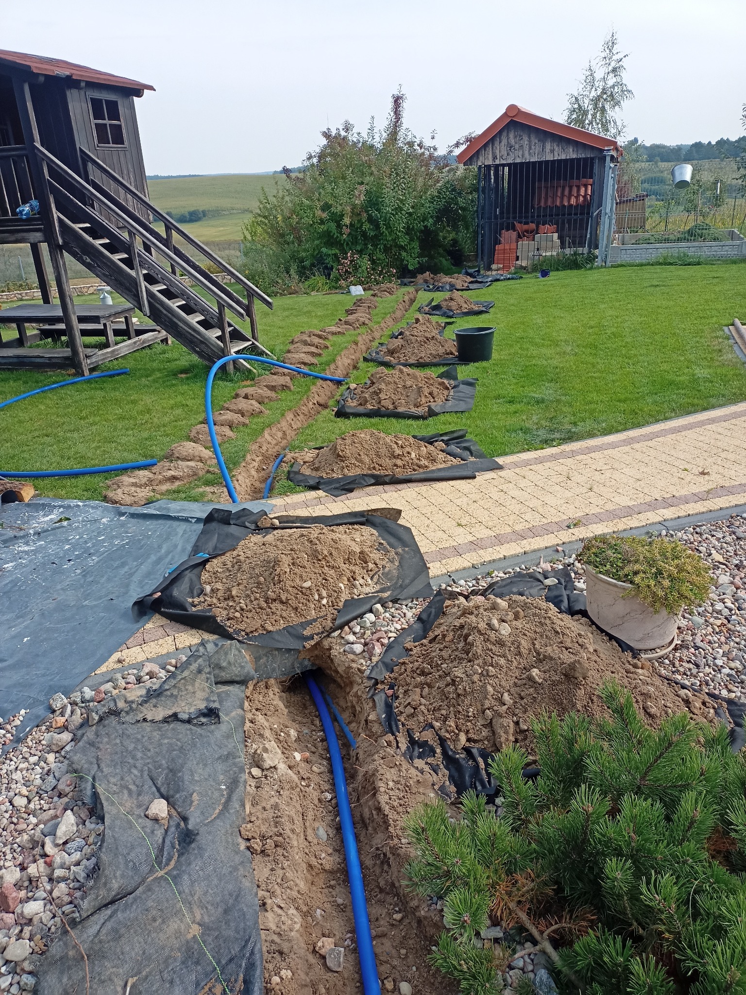 Wykopany rów w trawniku z ułożonymi niebieskimi rurami, przykryty czarną geowłókniną i usypanymi pryzmami ziemi, w tle domek na drzewie i budynek gospodarczy.