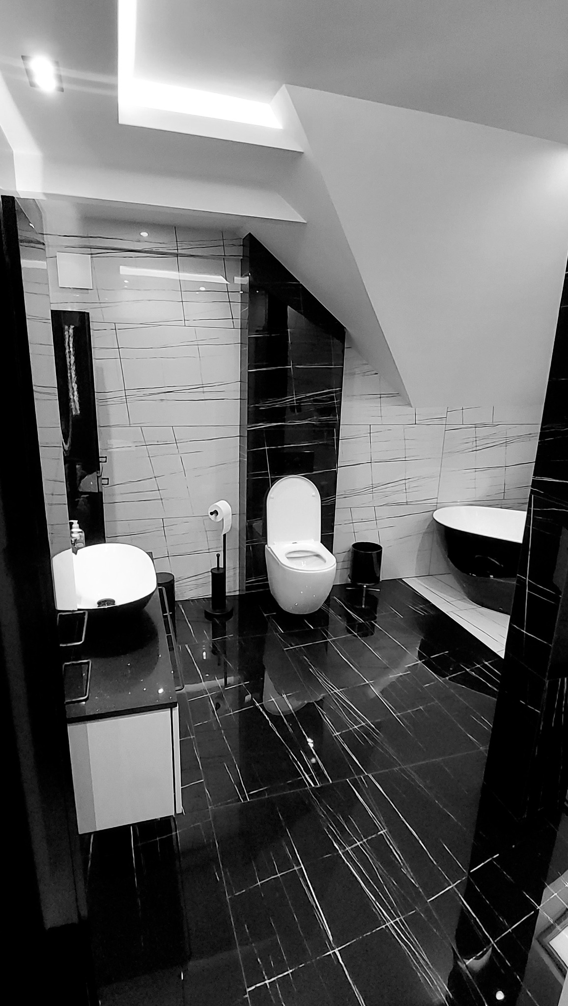 Monochromatyczna łazienka z czarno-białymi płytkami imitującymi marmur na podłodze i ścianach, z nowoczesną armaturą obejmującą umywalkę nablatową, toaletę i wannę wolnostojącą.