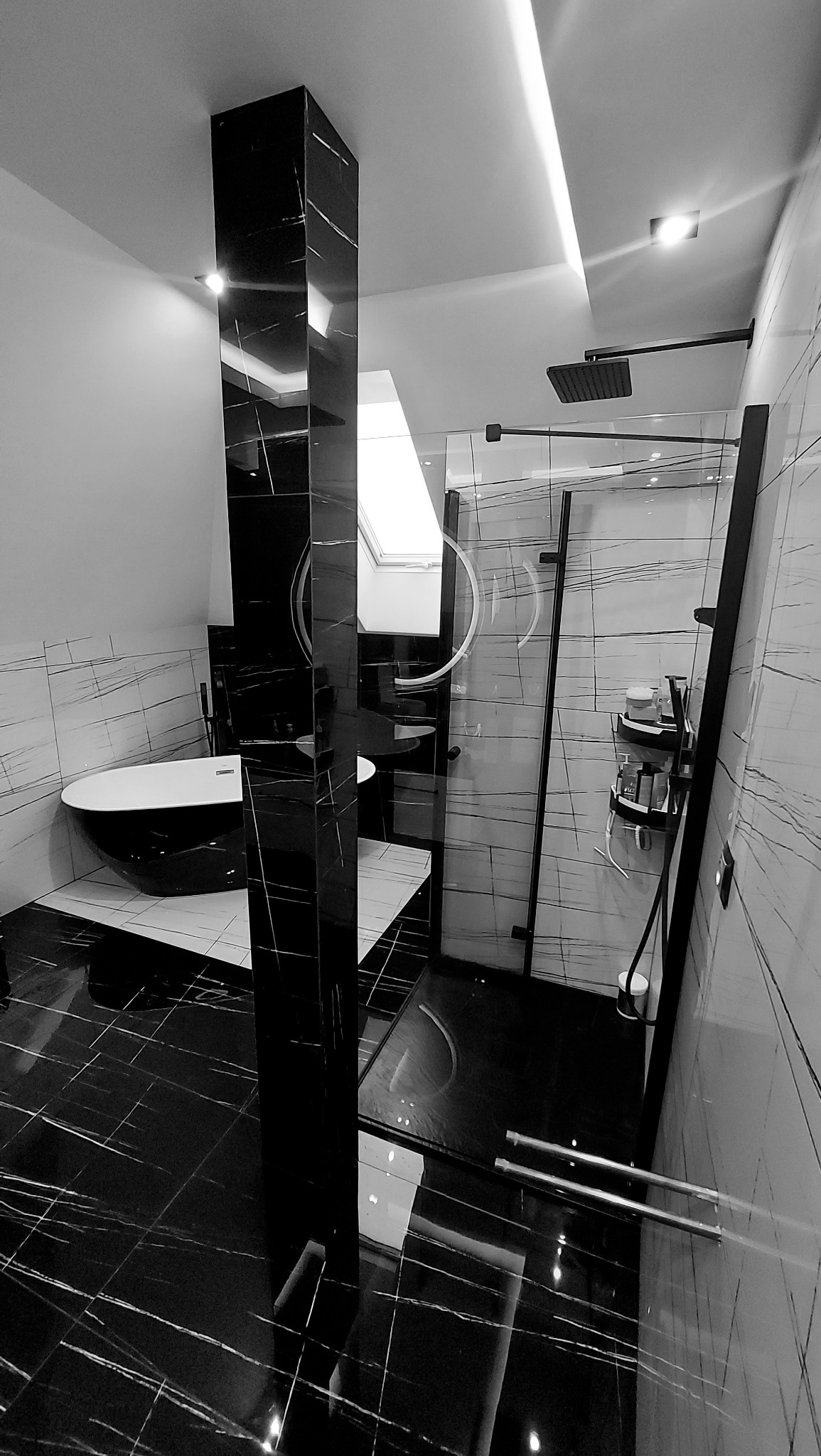 Wykończona łazienka w czerni i bieli z wolnostojącą wanną, kabiną prysznicową typu walk-in z czarnymi ramami i marmurowymi płytkami z białymi żyłami, oświetlona światłem dziennym z okna dachowego.