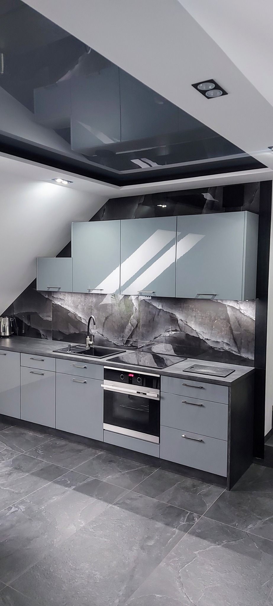 Nowoczesna kuchnia w odcieniach szarości: błyszczący sufit napinany, szafki z frontami na wysoki połysk, marmurowy backsplash i płytki podłogowe imitujące kamień.