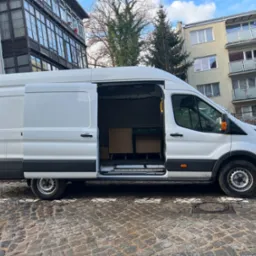 Biały bus z otwartymi drzwiami bocznymi, częściowo załadowany meblami, zaparkowany na brukowanej ulicy w miejskiej scenerii.