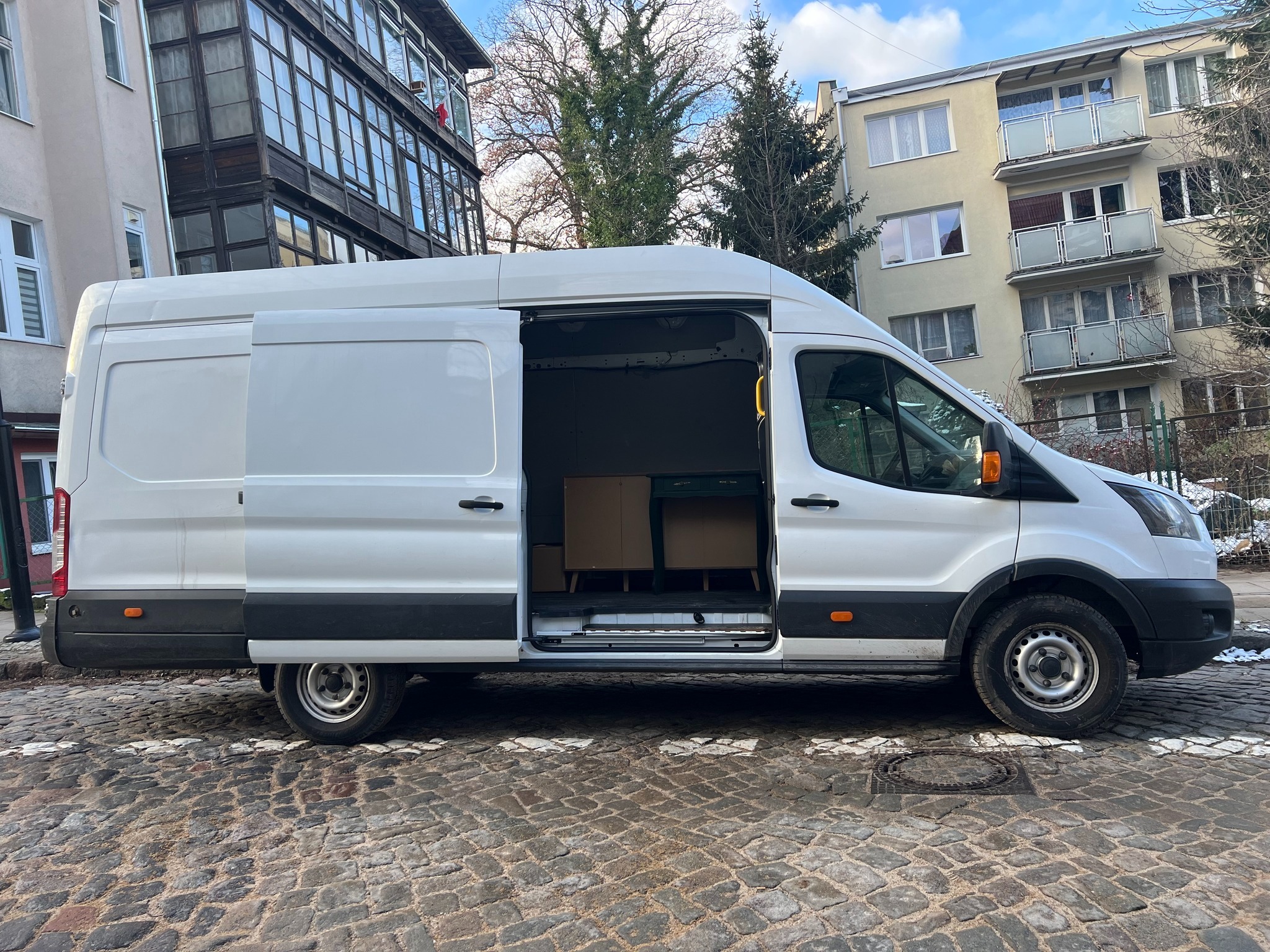 Biały bus z otwartymi drzwiami bocznymi, częściowo załadowany meblami, zaparkowany na brukowanej ulicy w miejskiej scenerii.