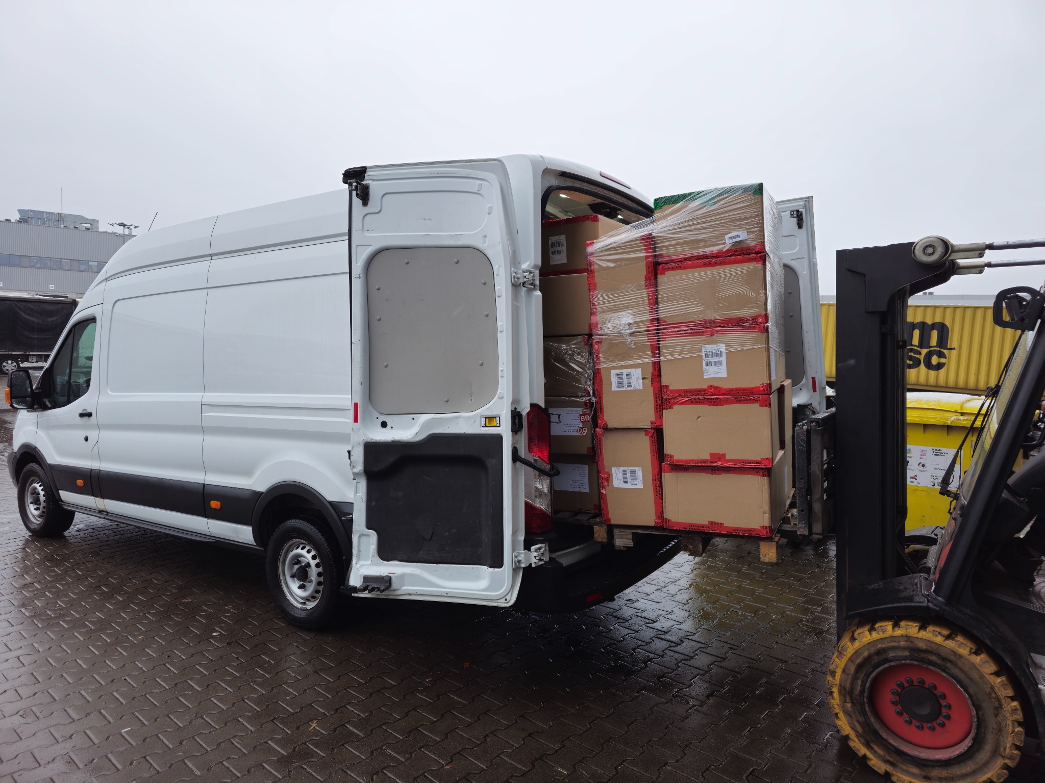 Biały bus z otwartymi drzwiami załadunkowymi, wypełniony kartonami zabezpieczonymi folią, podnoszony przez wózek widłowy na zewnątrz, na mokrym bruku.