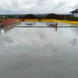 Świeżo wylany fundament domu jednorodzinnego z widocznym szalunkiem w kolorze żółtym i czerwonym, odbijającym pochmurne niebo. W tle dom w budowie i zieleń.