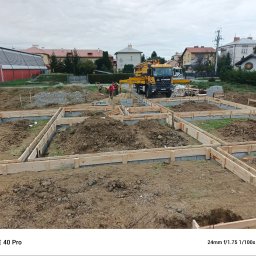 Krzysztof Wacek - Przygotowanie fundamentów pod budowę domu: drewniane szalunki, zbrojenie i betonowa pompa Styrobud w tle. Widoczni pracownicy w czerwonych kurtkach.