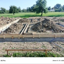 Krzysztof Wacek - Ziemia w wykopach fundamentowych zbrojonych stalą, otoczonych drewnianym szalunkiem na bloczkach betonowych, na tle zielonej łąki i drzew w słoneczny dzień.