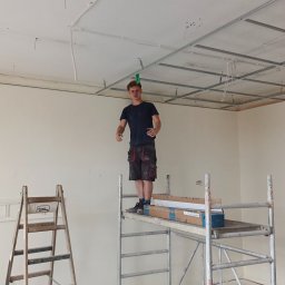 Krzysztof Wacek - Pracownik na rusztowaniu montuje metalowy stelaż sufitu podwieszanego w jasnym wnętrzu. Widoczna drabina, kable i częściowo zamontowana płyta.