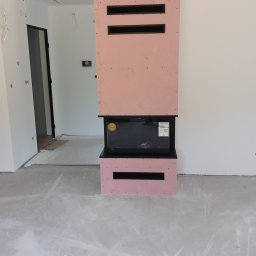 Krzysztof Wacek - Wnętrze z obudową kominka z płyty karton-gips w kolorze różowym, z czarnymi wnękami wentylacyjnymi. Podłoga szara, surowa. Widoczne niedokończone prace wykończeniowe.